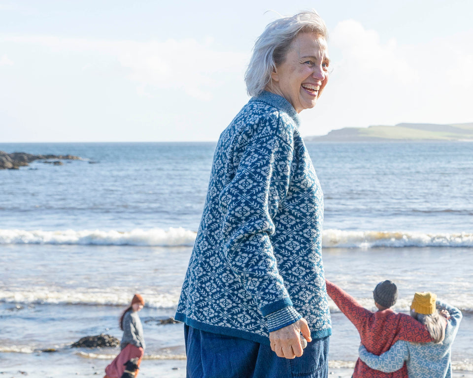 A woman with jaw length grey hair wears a blue and grey fair isle patterned jumper over a loose denim dress. She turns to face the camera. In the background, the sea at Hoswick stretches out towards Levenwick headland. On the shore a young woman with a black labrador and tow women with a rusty fair isle jumper and a pale blue and pale grey fair isle jumper are milling about. All three on the beach are wearing beanie hats.