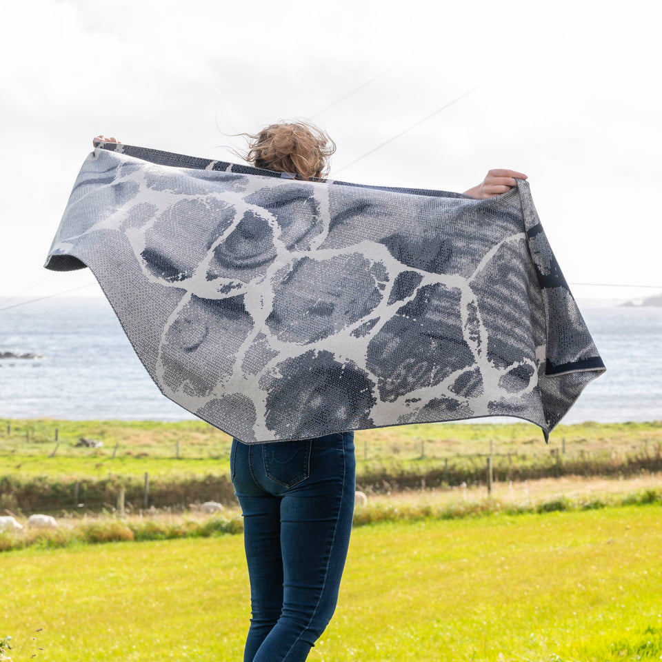 A woman with fair hair holds up a large, contemporary shawl with the paler side showing. In the background is the sea at Hoswick, Shetland