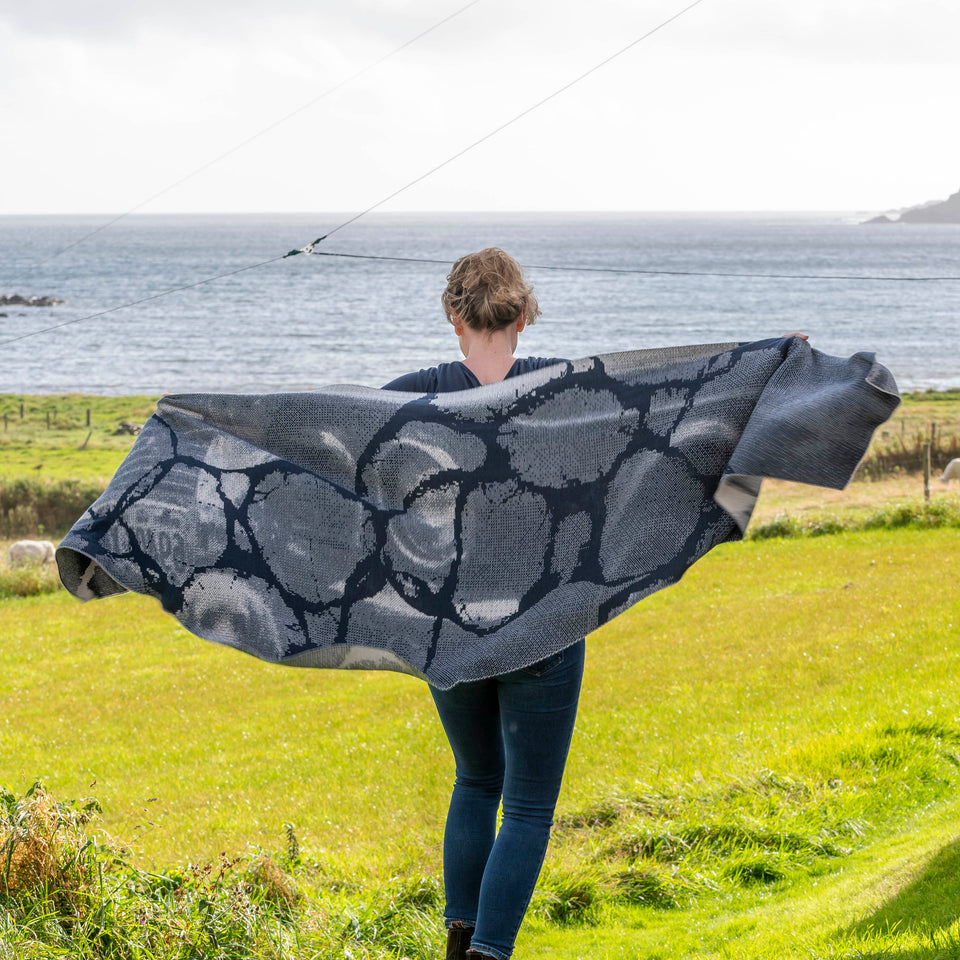 A woman with fair hair holds up a large, contemporary shawl with the darker side showing. In the background is the sea at Hoswick, Shetland
