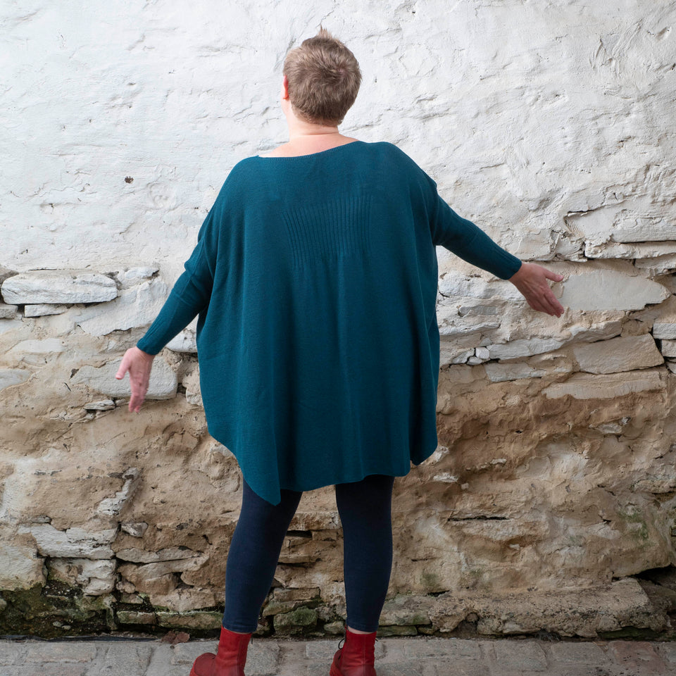 A white woman with short fair hair stands in a rustic stone shed. She has her back to the camera and is wearing a loose and finely knit jumper in dark teal over navy leggings and red chunky lace up boots. She holds her arms out to the sides to show the nipped in sleeves.