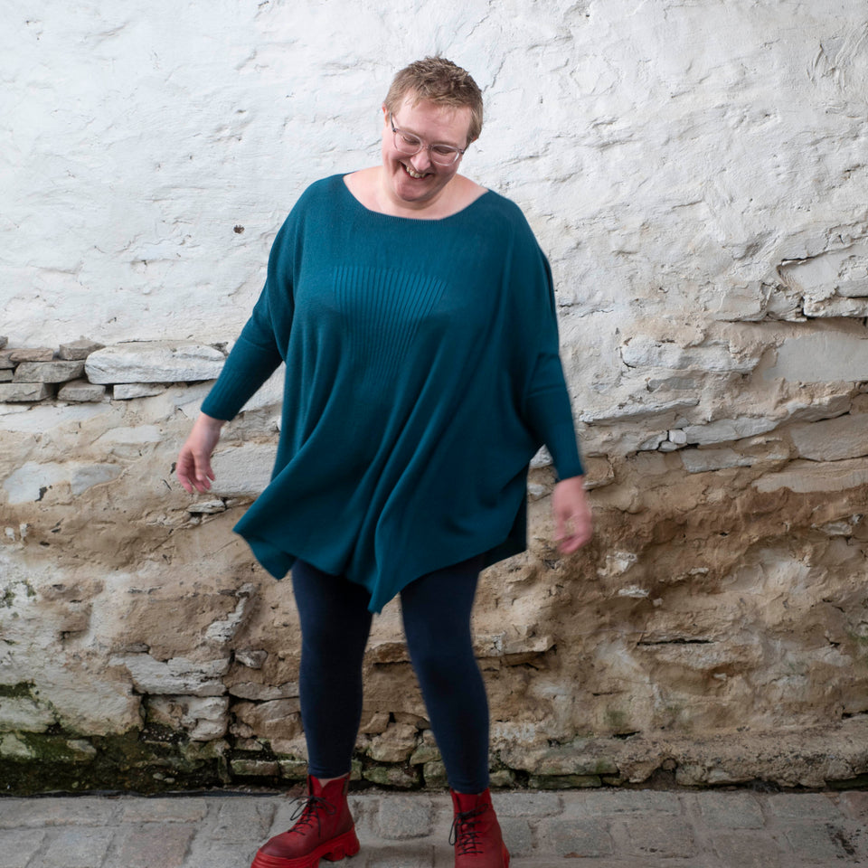 A white woman with short fair hair stands in a rustic stone shed. She faces the camera and looks down, smiling. She is wearing a loose and finely knit jumper in dark teal over navy leggings and red chunky lace up boots. She dances to show the movement in the sweater.