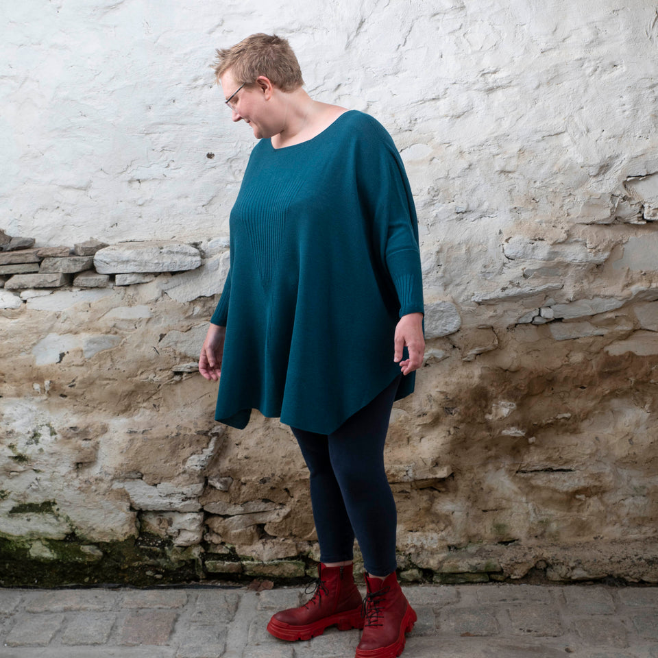 A white woman with short fair hair stands in a rustic stone shed. She stands side-on to the camera and looks slightly down. and is wearing a loose and finely knit jumper in dark teal over navy leggings and red chunky lace up boots.