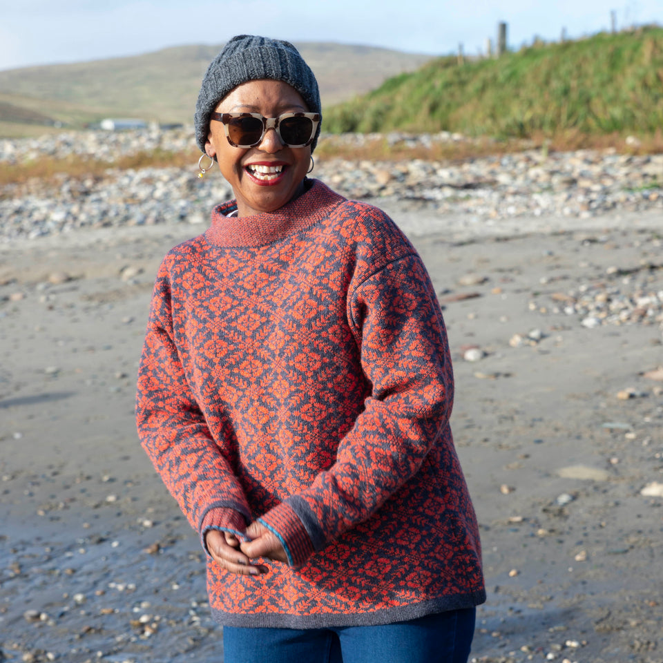 On the beach in Hoswick, a woman wears a patterned Fair Isle jumper in charcoal grey and rusty pinkish red. She is also wearing tortoisehell sunglasses and dark blue jeans, and a charcoal grey ribbed beanie.