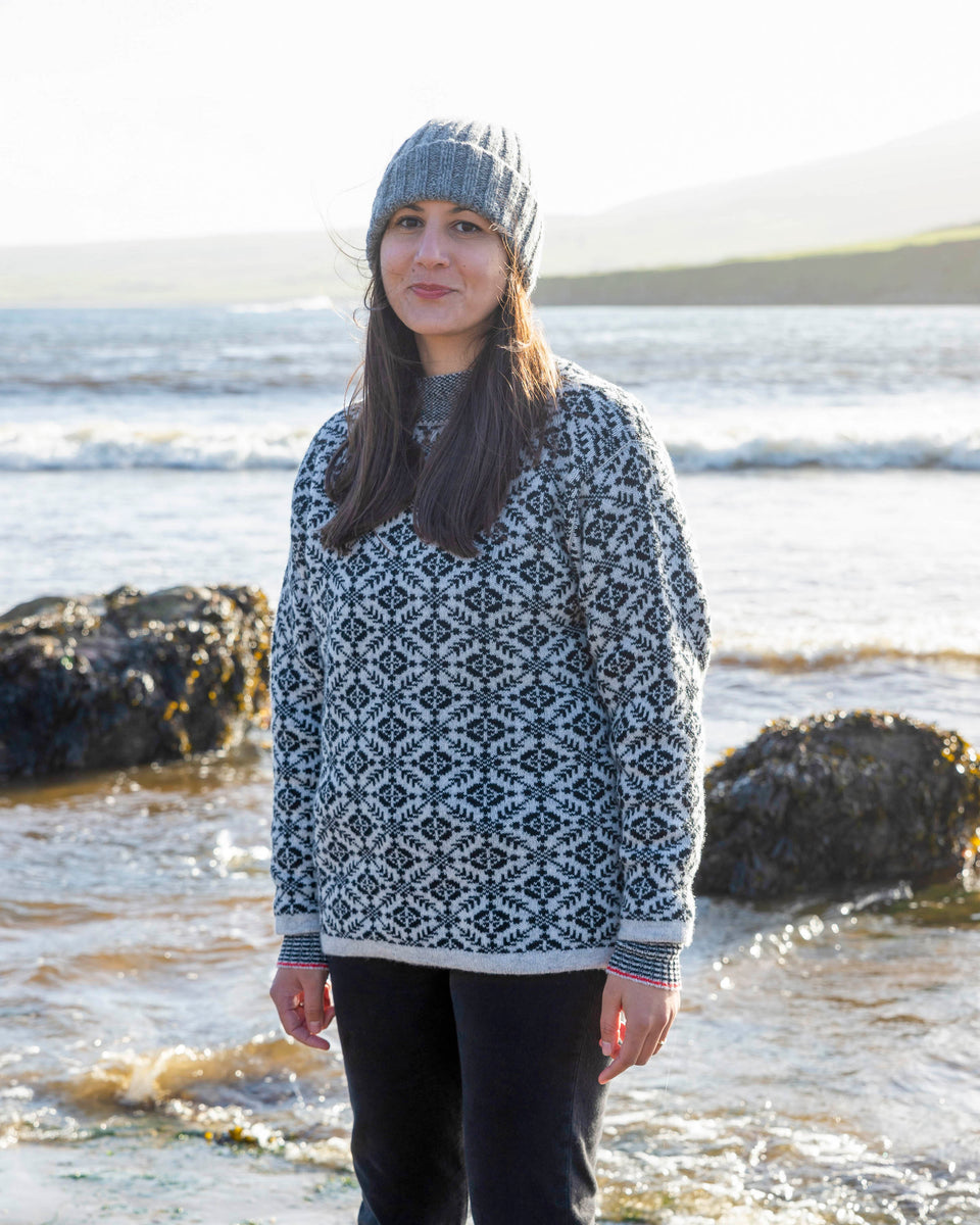 Fair Isle jumper in charcoal and pale grey with red stripe at cuff. A woman with olive skin and long dark hair stands on the beach at Hoswick, Shetland. She is wearing a chunky grey beanie hat, the Fair Isle jumper in charcoal and pale grey and black jeans.