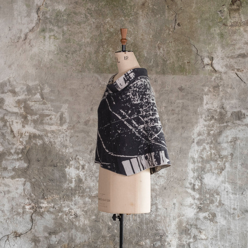 Knitted Byre capelet. Abstract, graphic design in an asymmetric shape. Shown in Charcoal and stone white. Shown on mannequin from side, with non-opening side showing.