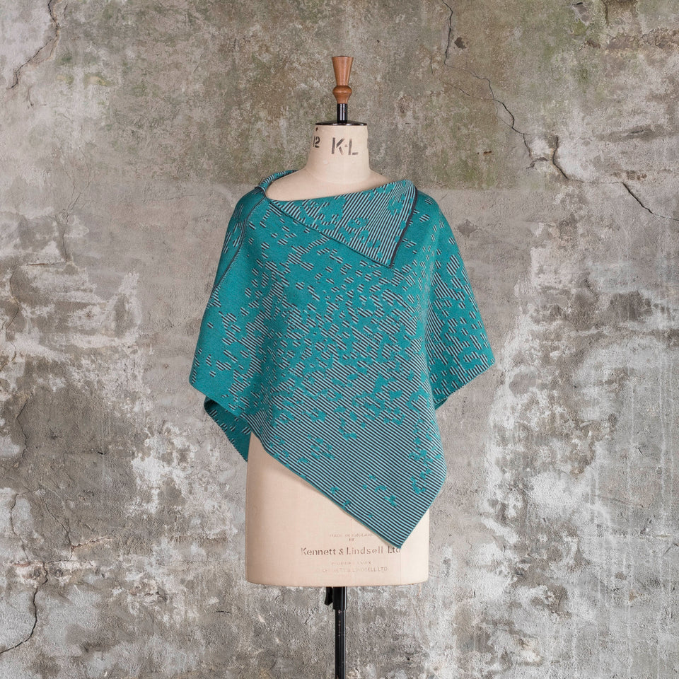 On a vintage mannequin against a rustic, old plastered wall, is a finely knitted cape with a mottled and striped pattern. There are two teals, one light and the other dark, with a very dark blue fine stripe. Shown from the front.