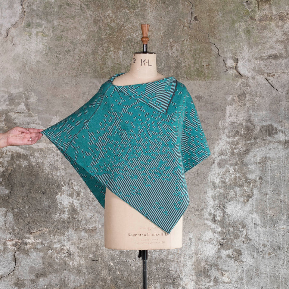 On a vintage mannequin against a rustic, old plastered wall, is a finely knitted cape with a mottled and striped pattern. There are two teals, one light and the other dark, with a very dark blue fine stripe. Shown from the front with one side pulled out by a hand just in shot.
