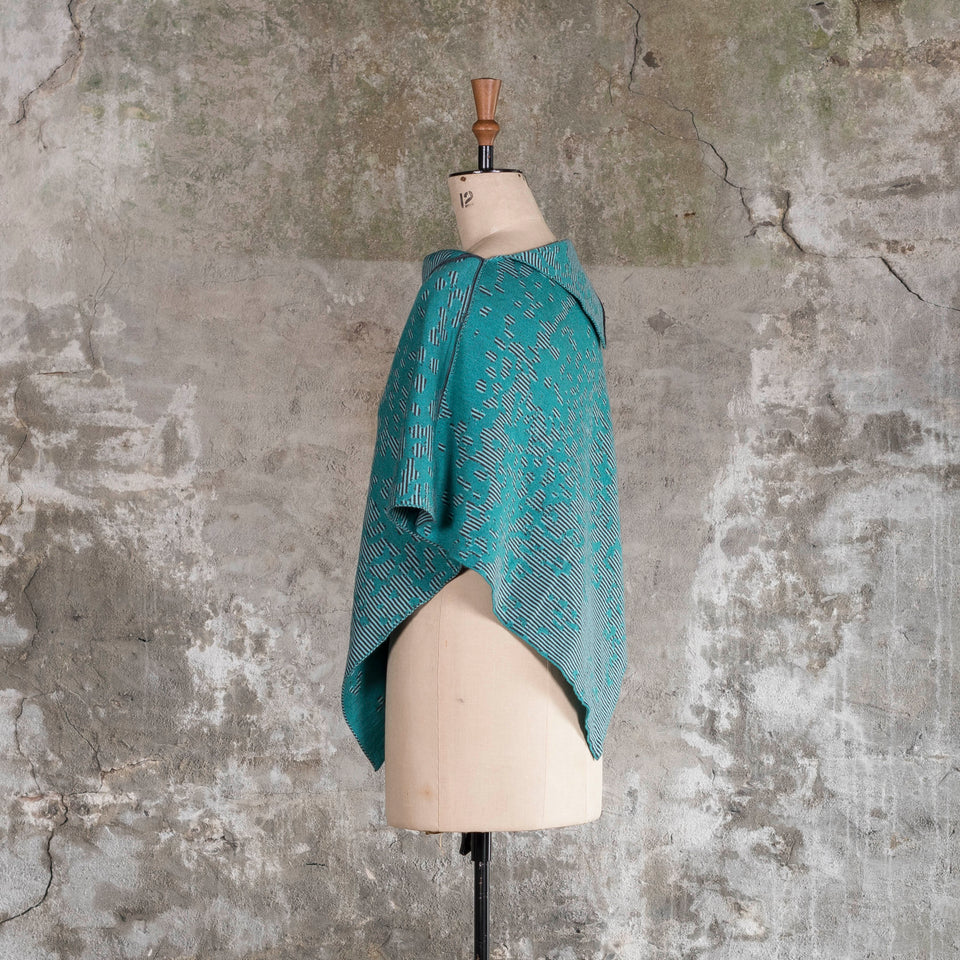 On a vintage mannequin against a rustic, old plastered wall, is a finely knitted cape with a mottled and striped pattern. There are two teals, one light and the other dark, with a very dark blue fine stripe. Shown from the left side