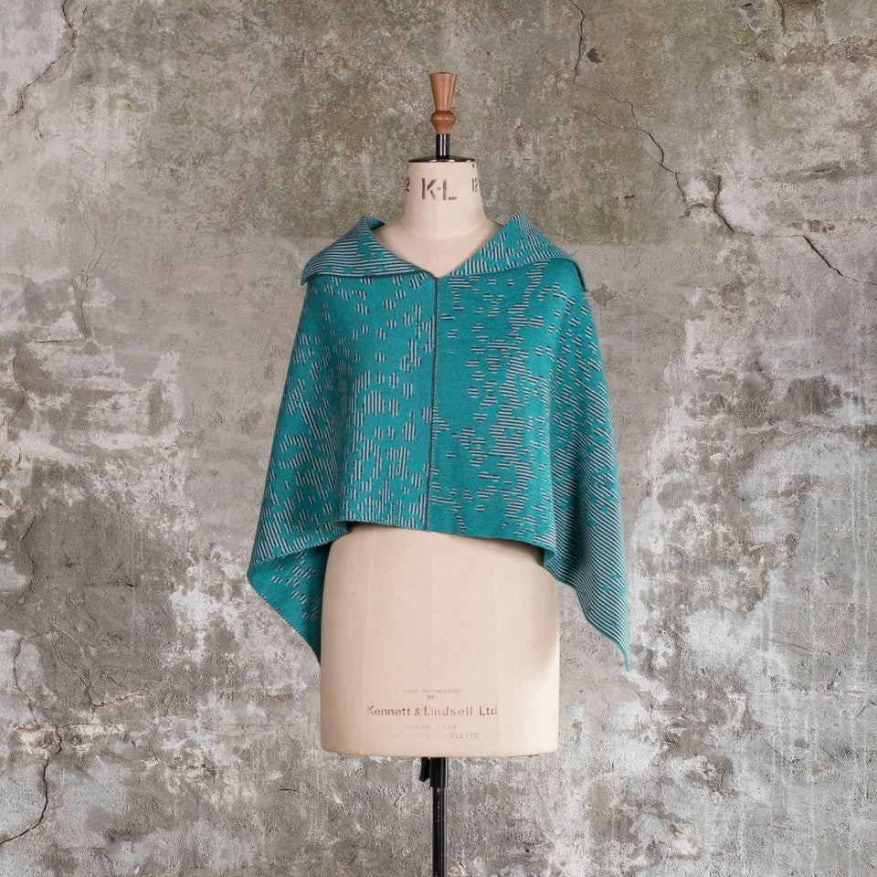 On a vintage mannequin against a rustic, old plastered wall, is a finely knitted cape with a mottled and striped pattern. There are two teals, one light and the other dark, with a very dark blue fine stripe. Shown from the front with the cape worn straight - with long edges at the sides and collar folded out.