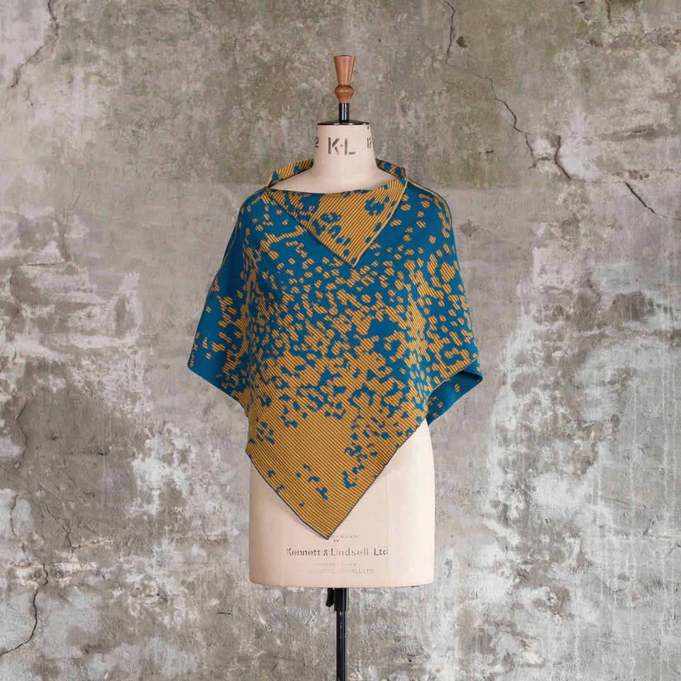 On a vintage mannequin against a rustic, old plastered wall, is a finely knitted cape with a mottled and striped pattern. The colours are very dark teal blue with ochre yellow, one light and the other darker. The yellow areas are striped with the two gold ochres. Shown from the front with the cape worn with the longest edge at front and back.