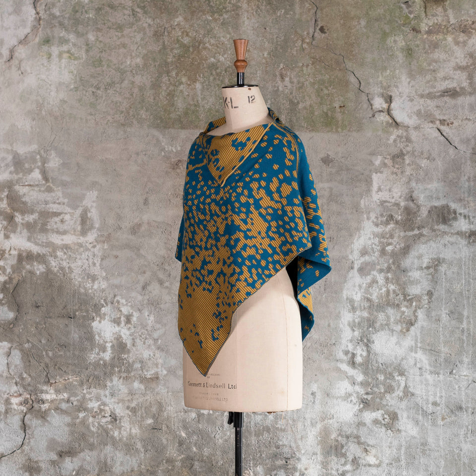 On a vintage mannequin against a rustic, old plastered wall, is a finely knitted cape with a mottled and striped pattern. The colours are very dark teal blue with ochre yellow, one light and the other darker. The yellow areas are striped with the two gold ochres. Shown from the left side, turned three-quarters,with the cape worn with the longest edge at front and back.