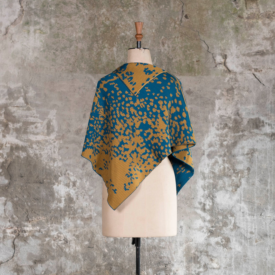 On a vintage mannequin against a rustic, old plastered wall, is a finely knitted cape with a mottled and striped pattern. The colours are very dark teal blue with ochre yellow, one light and the other darker. The yellow areas are striped with the two gold ochres. Shown from the back with the cape worn with the longest edge at front and back.