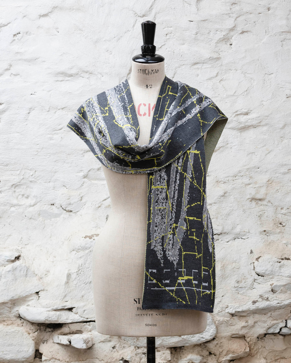 A grey scarf with abstract patterning and linear limey yellow  highlights. Shown loosely draped with one end over shoulder. Large and luxurious scarf knitted in fine merino. Shown on a vintage mannequin against a rustic whitewashed wall