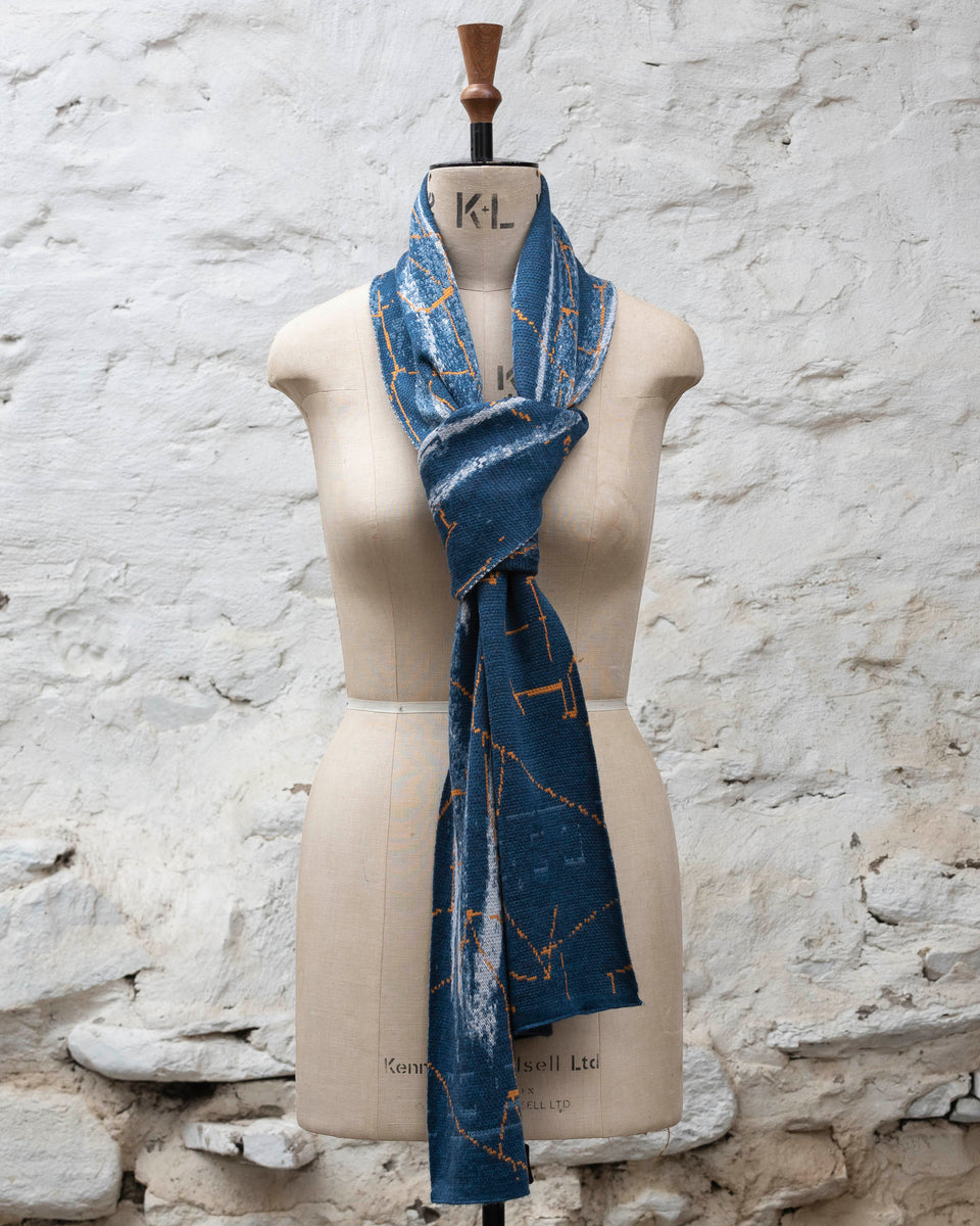 A blue scarf with abstract patterning and linear orange highlights. Shown tied in a tie knot. Large and luxurious scarf knitted in fine merino. Shown on a vintage mannequin against a rustic whitewashed wall