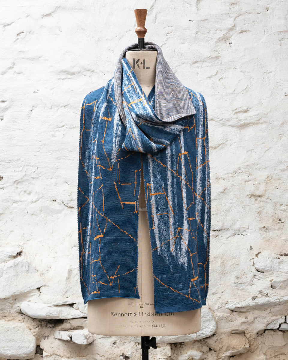 A blue scarf with abstract patterning and linear orange highlights. Shown loosely wrapped with even ends. Large and luxurious scarf knitted in fine merino. Shown on a vintage mannequin against a rustic whitewashed wall
