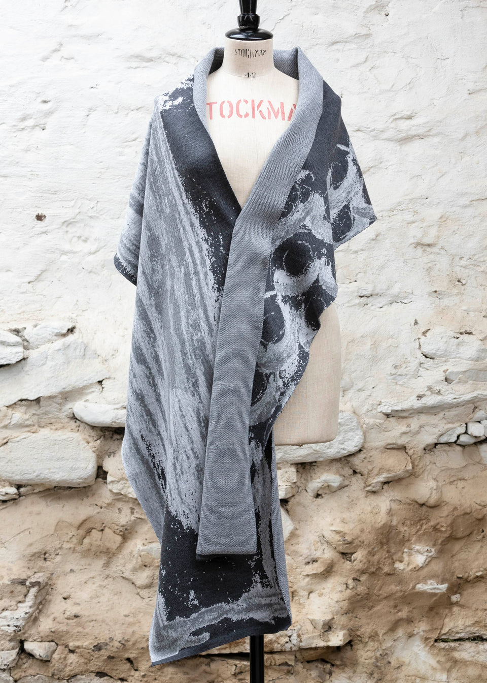 Finely knitted shawl in luxurious merino, made in Scotland. Shown on a vintage mannequin against a whitewashed wall. Abstract design in sea greys with curvilinear motifs. Draped to show whole shawl from the front.