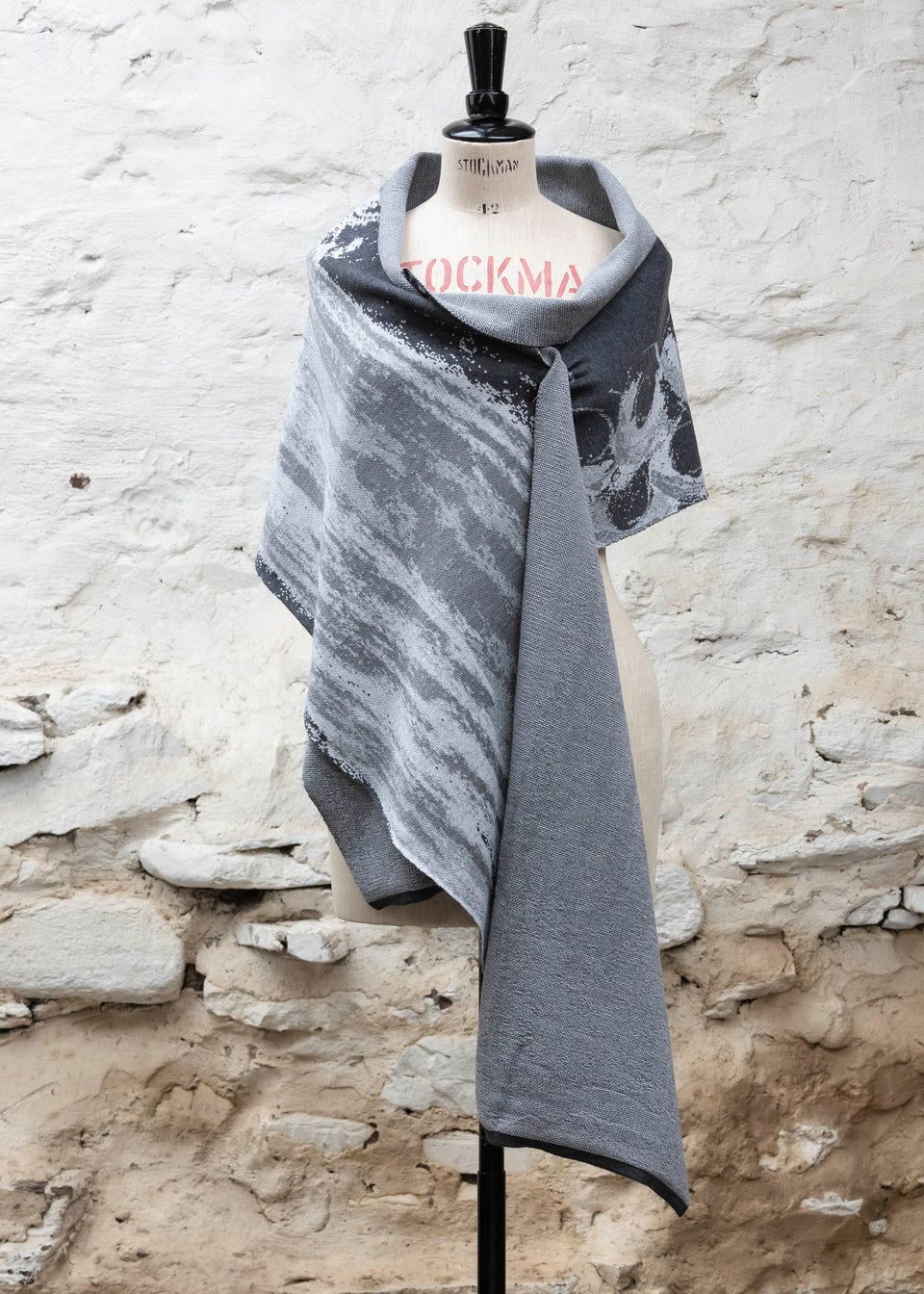 Finely knitted shawl in luxurious merino, made in Scotland. Shown on a vintage mannequin against a whitewashed wall. Abstract design in sea greys with curvilinear motifs. Draped to show whole shawl from the front, with a pin