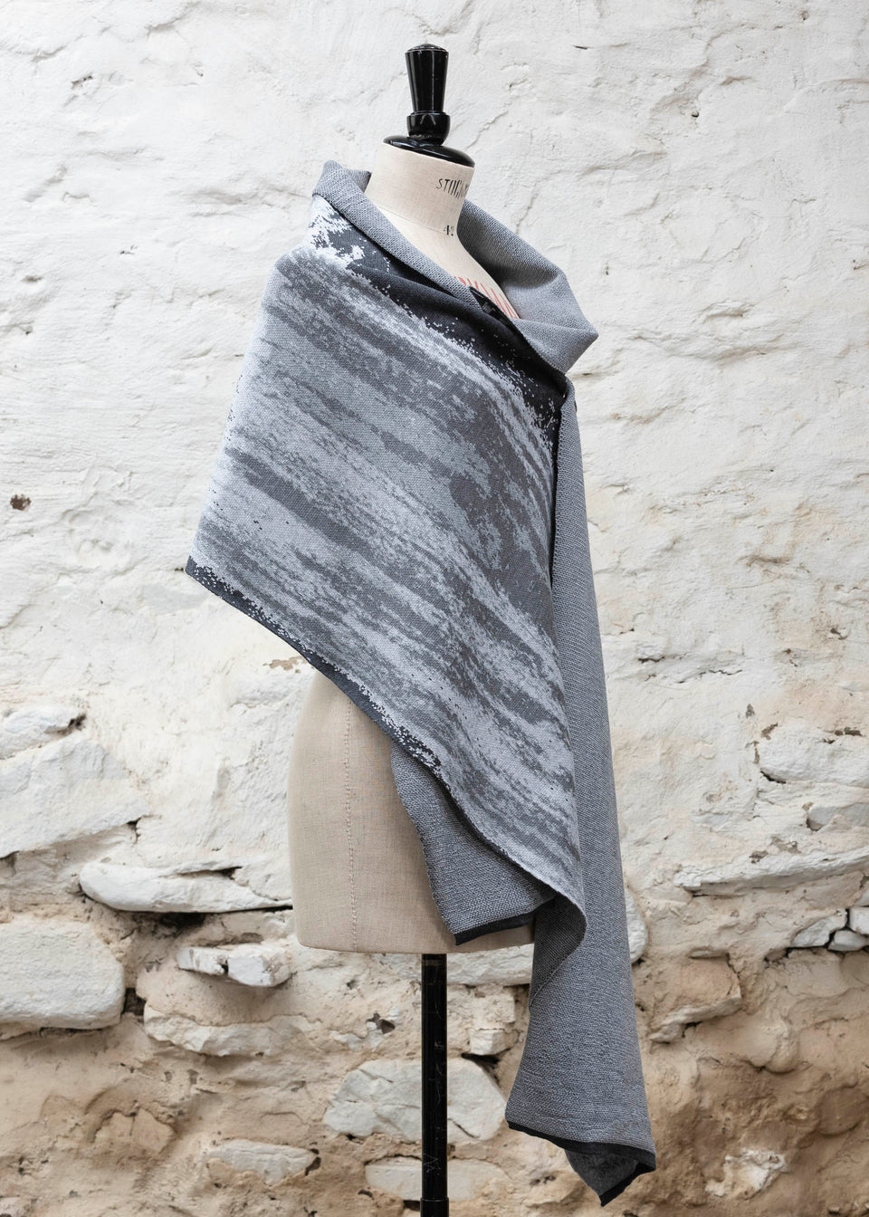 Finely knitted shawl in luxurious merino, made in Scotland. Shown on a vintage mannequin against a whitewashed wall. Abstract design in sea greys with curvilinear motifs. Draped to show shawl pinned from side
