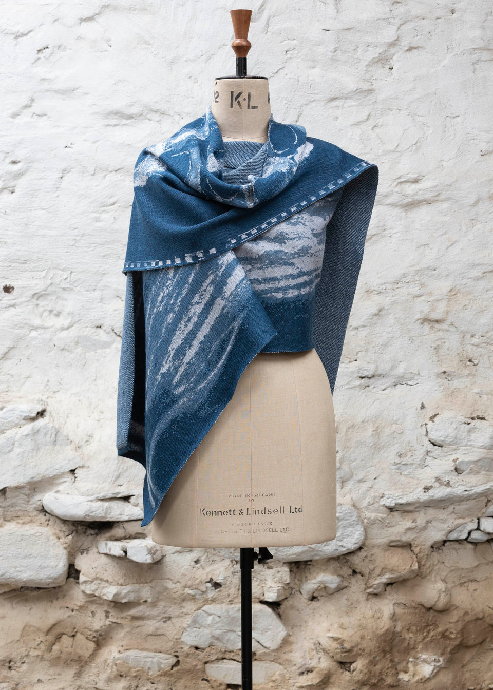 Finely knitted shawl in luxurious merino, made in Scotland. Shown on a vintage mannequin against a whitewashed wall. Abstract design in sea blues with curvilinear motifs. Wrapped around for a shorter style with a pin and shown from front