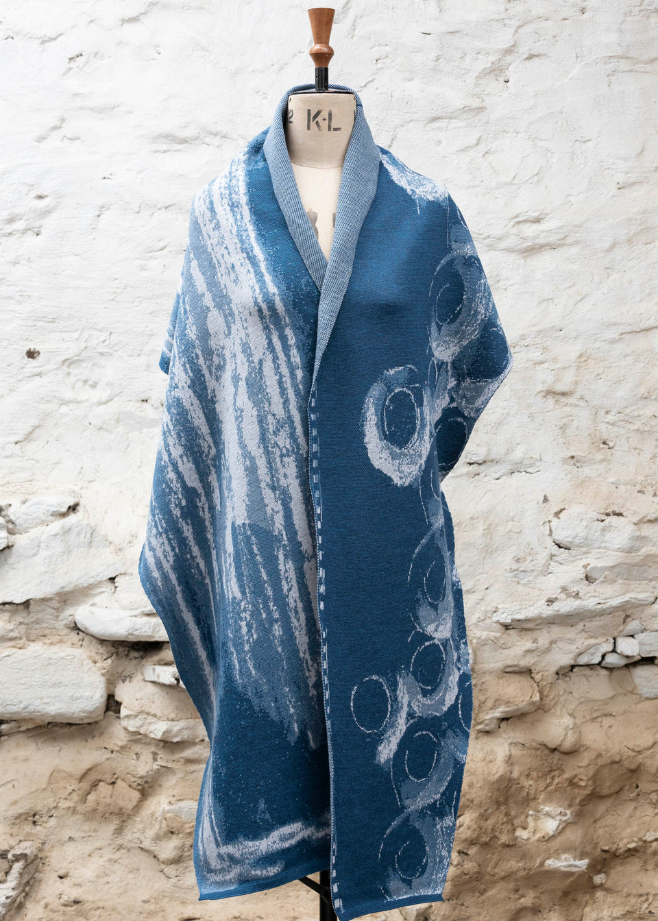 Finely knitted shawl in luxurious merino, made in Scotland. Shown on a vintage mannequin against a whitewashed wall. Abstract design in sea blues with curvilinear motifs. Draped to show full wrap from the front