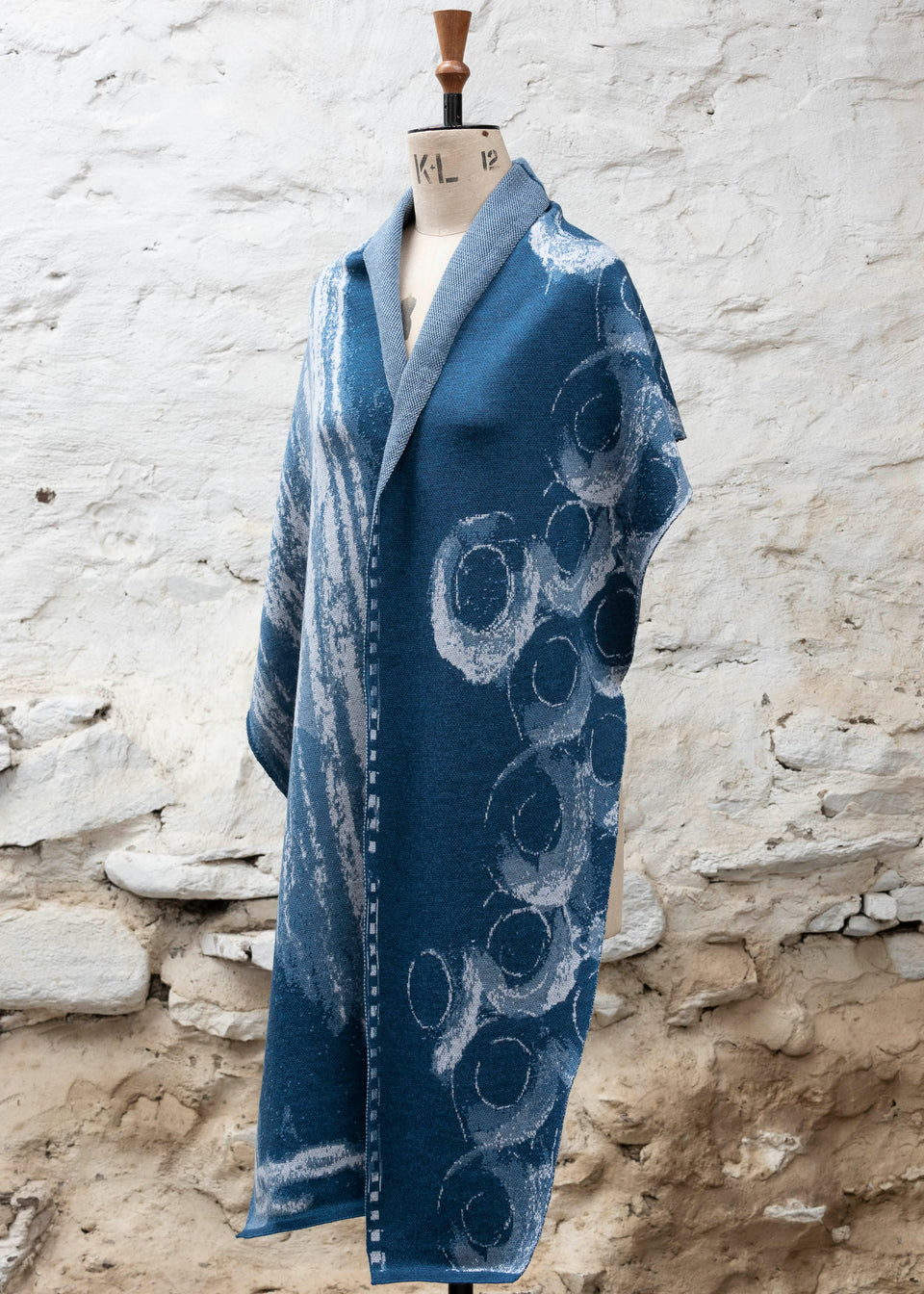 Finely knitted shawl in luxurious merino, made in Scotland. Shown on a vintage mannequin against a whitewashed wall. Abstract design in sea blues with curvilinear motifs. Draped to show full wrap from the side