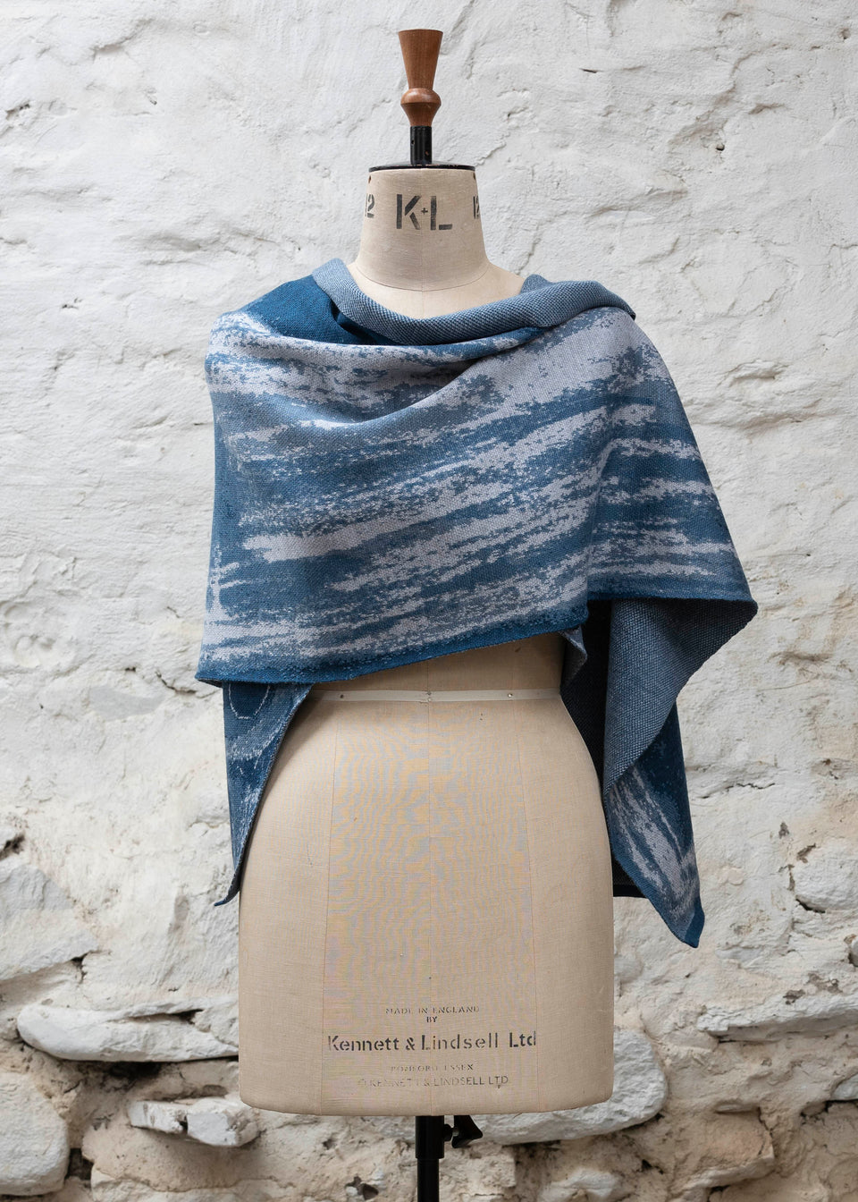 Finely knitted shawl in luxurious merino, made in Scotland. Shown on a vintage mannequin against a whitewashed wall. Abstract design in sea blues with curvilinear motifs. Wrapped around for a shorter style and shown from front