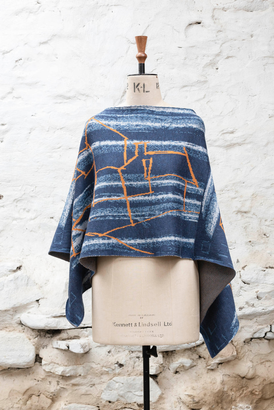 A knitted poncho, made in Shetland in a contemporary design by Nielanell with abstract blues and orange linear marks. Shown front on, with sides flowing down, on a vintage mannequin against a rustic white washed wall