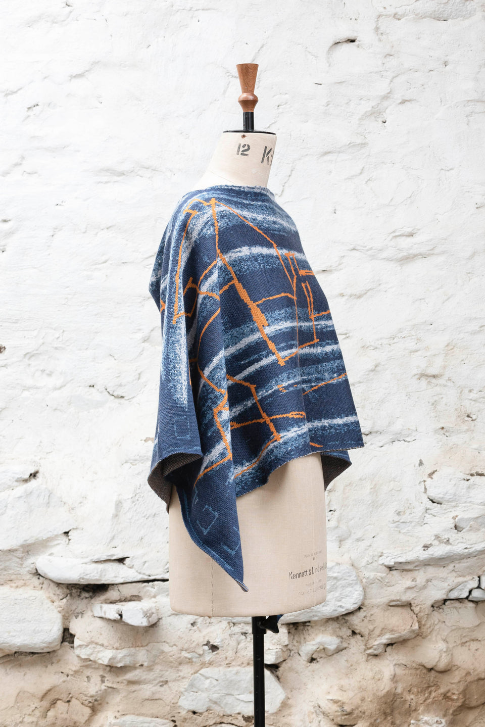 A knitted cape, made in Shetland in a contemporary design by Nielanell with abstract blues and orange linear marks. Shown side on, on a vintage mannequin against a rustic white washed wall