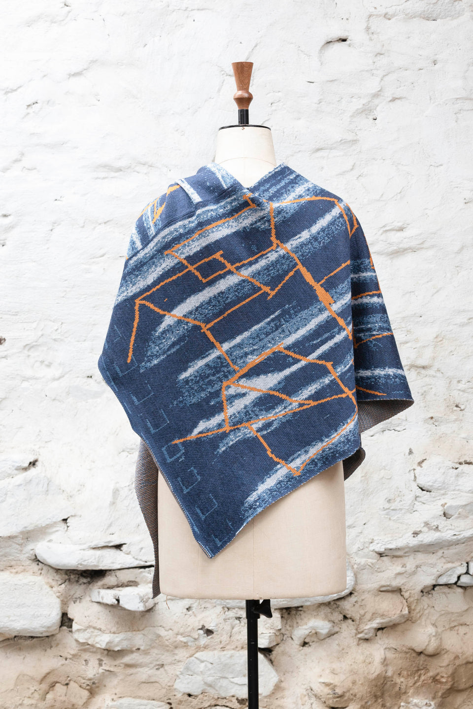 A knitted poncho, made in Shetland in a contemporary design by Nielanell with abstract blues and orange linear marks. Shown from the back, and with the neck arranged as a V, on a vintage mannequin against a rustic white washed wall