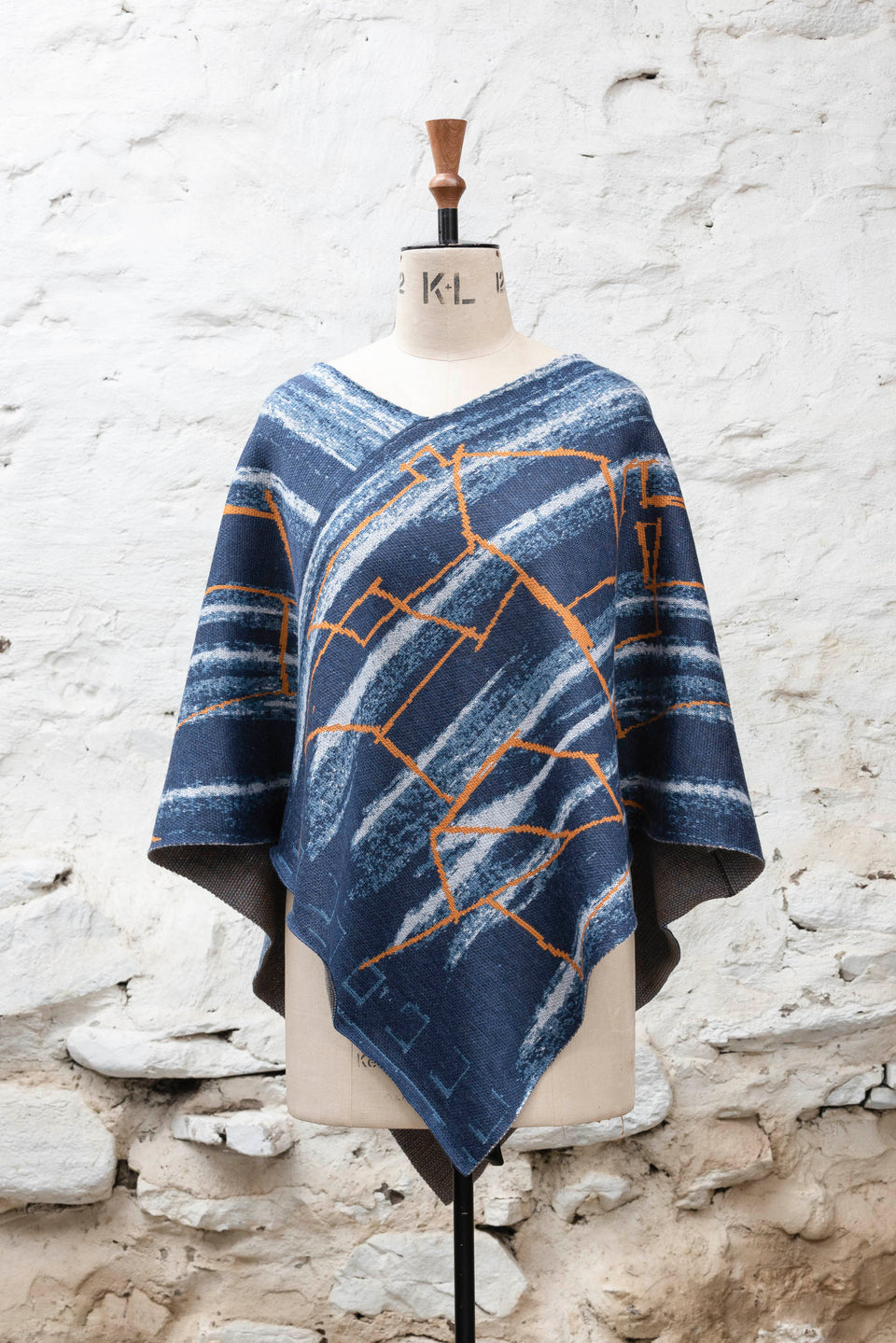 A knitted poncho, made in Shetland in a contemporary design by Nielanell with abstract blues and orange linear marks. Shown front on, and with the neck arrange as a V, on a vintage mannequin against a rustic white washed wall