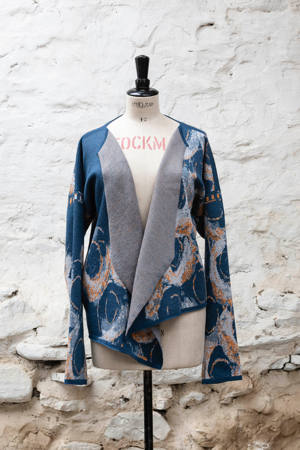 Knitted jacket inspired by the Shetland coastal village of Hoswick. Abstract curvilinear motif in blues with orange highlights . Asymmetric design with lapels that fold back. Shown front-on, on a vintage mannequin, against a white washed rustic stone wall