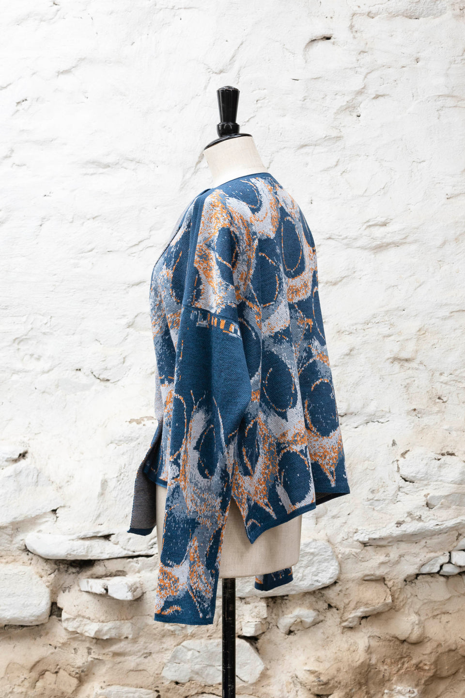 Knitted jacket inspired by the Shetland coastal village of Hoswick. Abstract curvilinear motif in blues with orange highlights . Asymmetric design with lapels that fold back. Shown side-on, on a vintage mannequin, against a white washed rustic stone wall