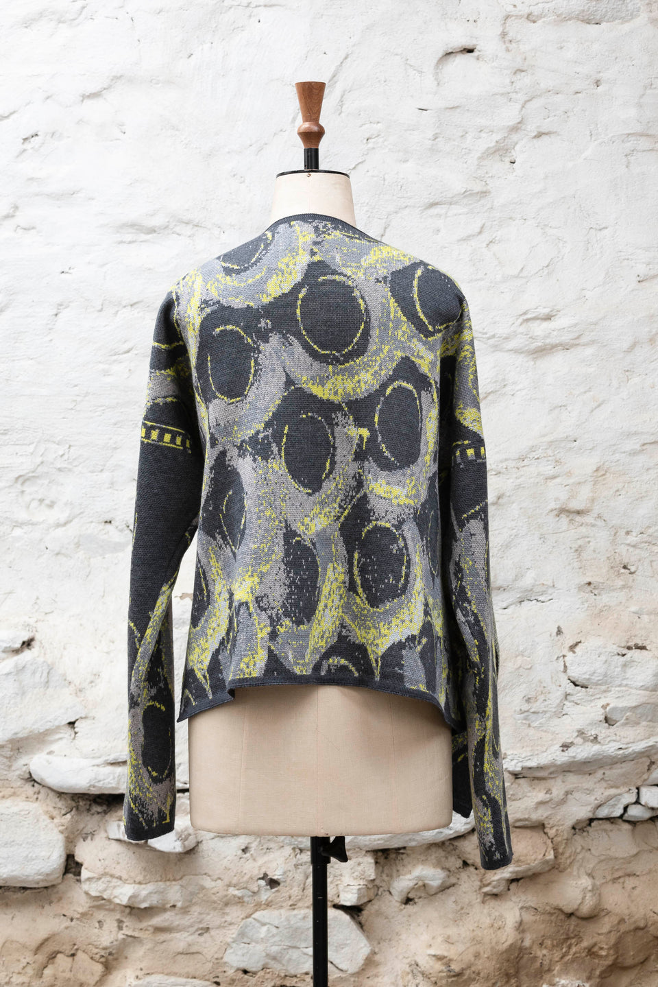Knitted jacket inspired by the Shetland coastal village of Hoswick. Abstract curvilinear motif in greys and fluoro yellow. Asymmetric design with lapels that fold back. Shown from the back, on a vintage mannequin, against a white washed rustic stone wall