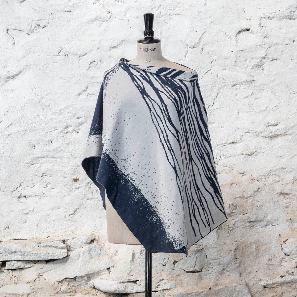 On a vintage dressmakers mannequin shown against a rustic whitewashed wall, a finely knitted poncho in a dark blue and off white. The pattern has an abstract, wavy linear design across one section of the cape. Shown from the front.