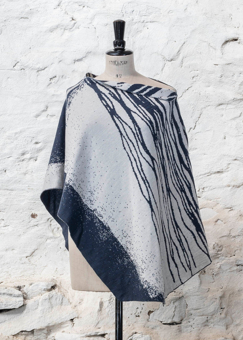 On a vintage dressmakers mannequin shown against a rustic whitewashed wall, a finely knitted poncho in a dark blue and off white. The pattern has an abstract, wavy linear design across one section of the cape. Shown from the front.