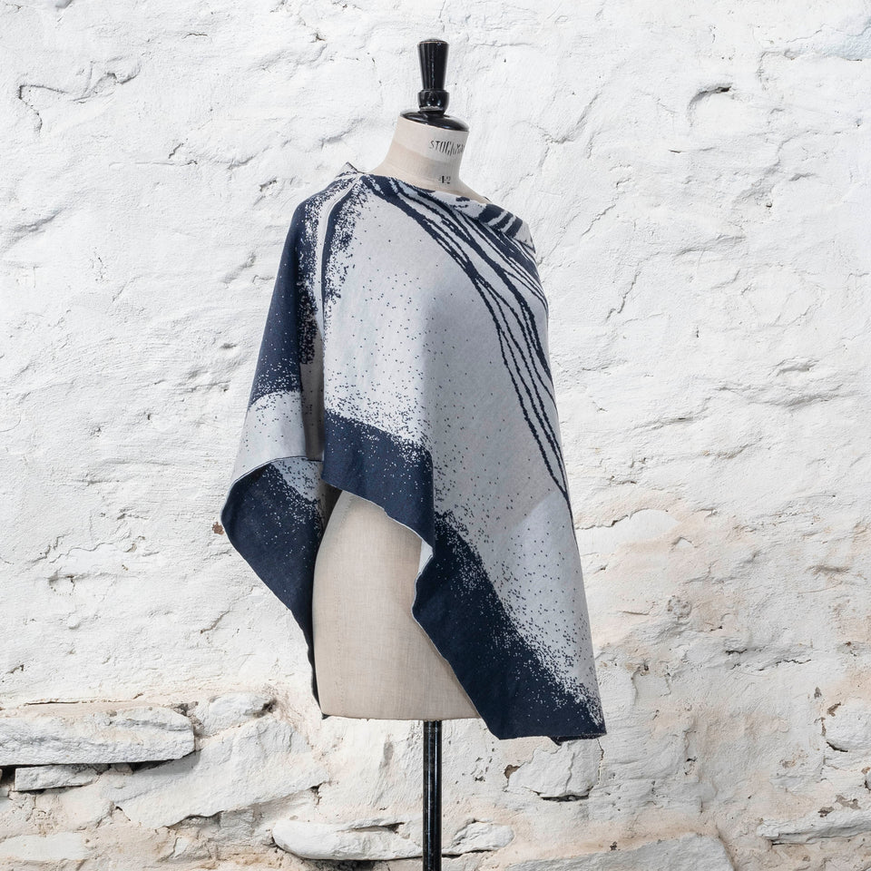 On a vintage dressmakers mannequin shown against a rustic whitewashed wall, a finely knitted poncho in a dark blue and off white. The pattern has an abstract, wavy linear design across one section of the cape. Shown from the left side, with panels of specked blue and white visible.