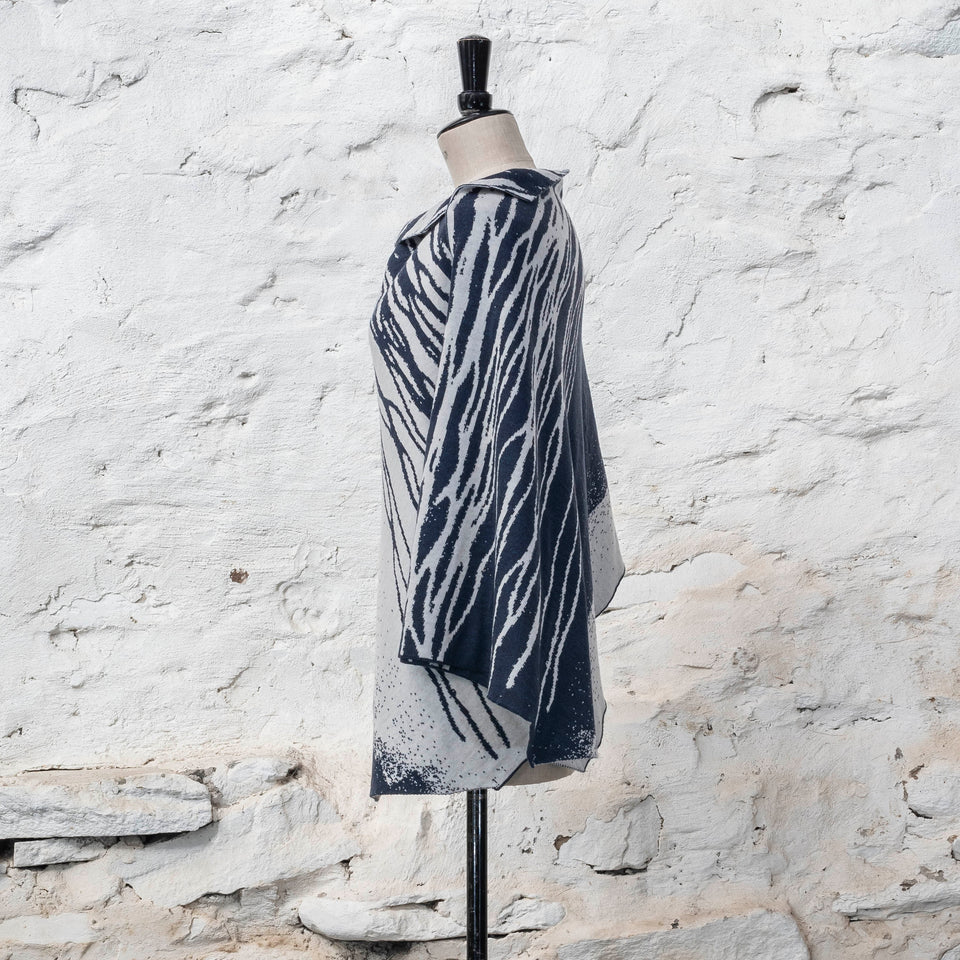 On a vintage dressmakers mannequin shown against a rustic whitewashed wall, a finely knitted poncho in a dark blue and off white. The pattern has an abstract, wavy linear design across one section of the cape. Shown from the right. The linear pattern is shown with both sections reversed out at the shoulder. Blue on white and white on blue.