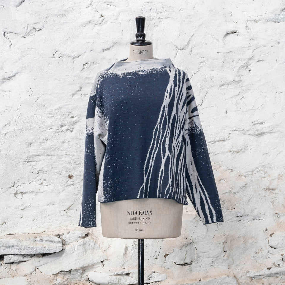 Contemporary jumper on a vintage mannequin against a rustic whitewashed wall, a finely knitted merino jumper in double faced jacquard. A linear design flows in waves down one side of the jumper - here on the front off-white against  deep midnight blue (the jumper has been turned around). There are areas of white with speckle and blue areas with white speckle.  The jumper  is shown from the front and has a stand up neckline.