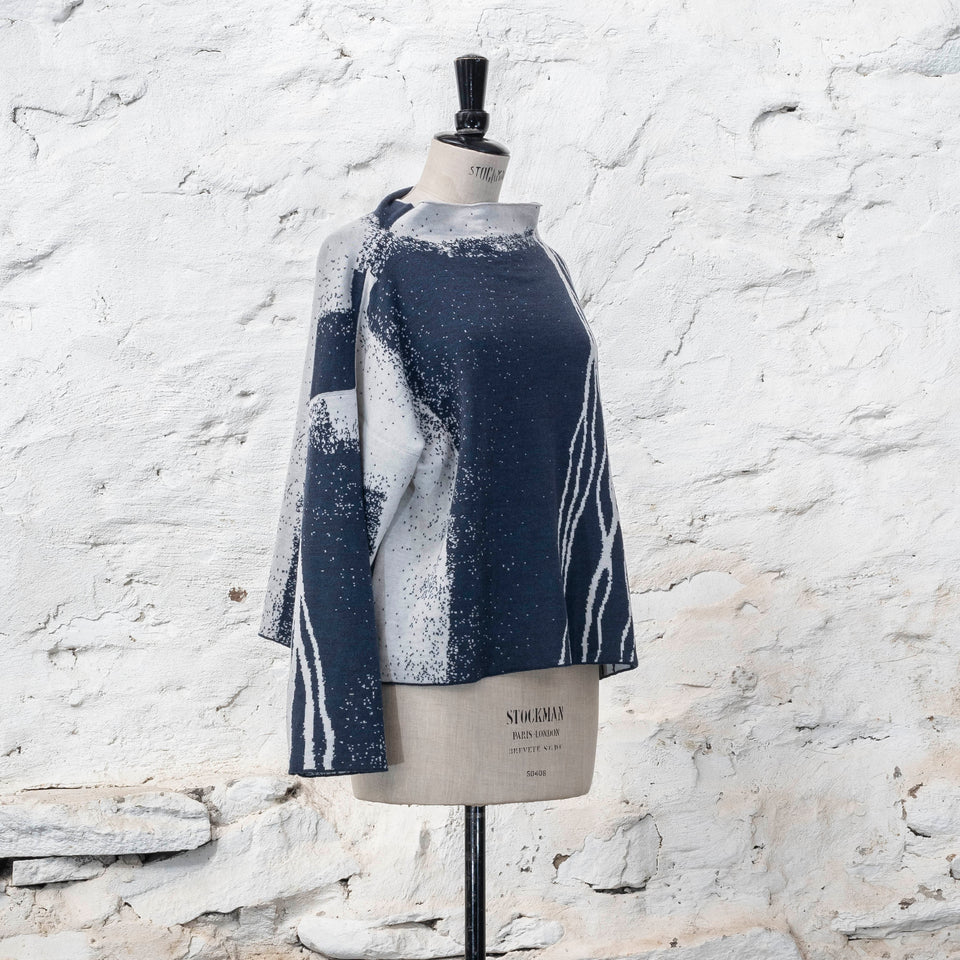 on a vintage mannequin against a rustic whitewashed wall, a finely knitted merino jumper in double faced jacquard. A linear design flows in waves down one side of the jumper - here on the front off-white against  deep midnight blue (the jumper has been turned around). There are areas of white with speckle and blue areas with white speckle.  The jumper  is shown from the right at a three quarter angle and has a stand up neckline.