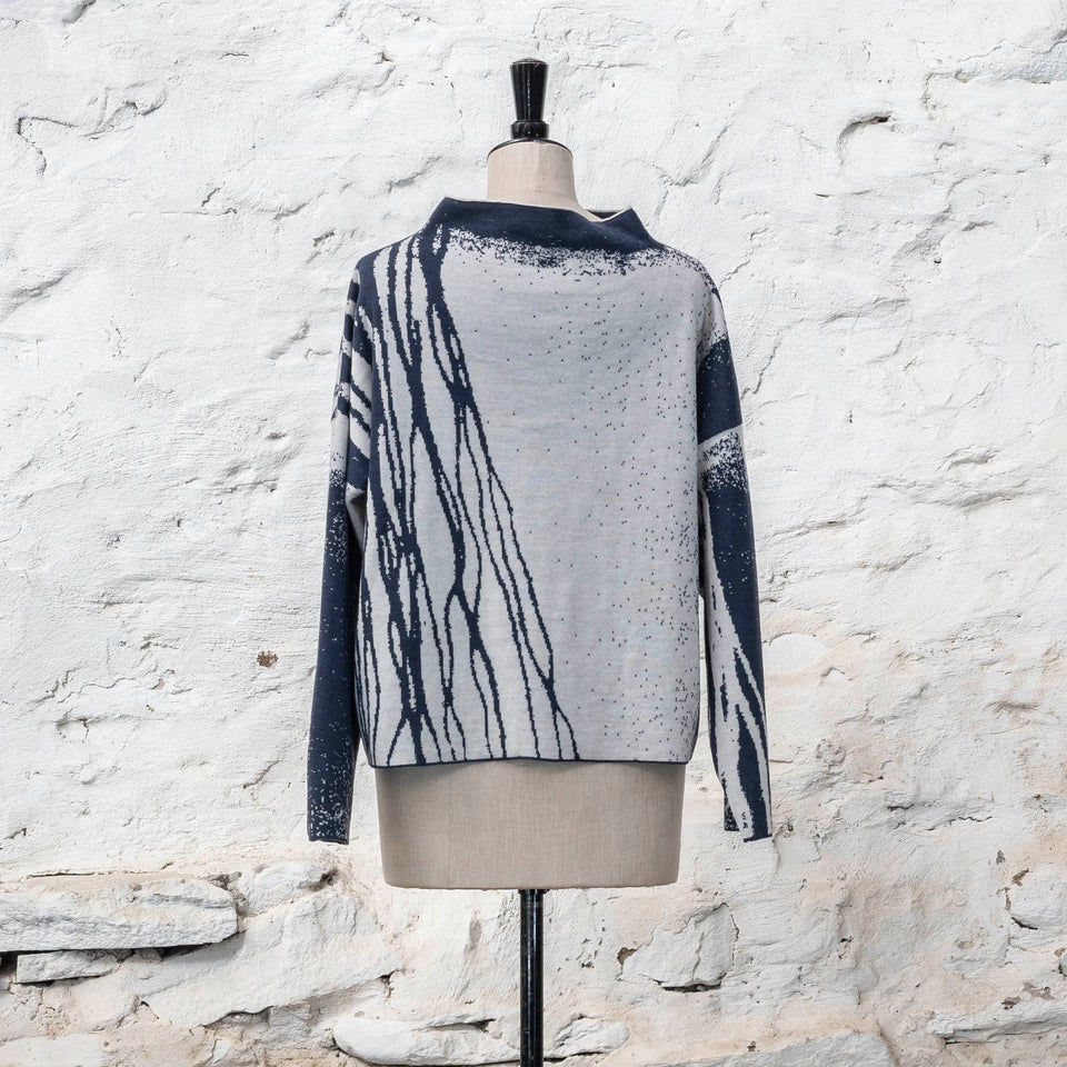 on a vintage mannequin against a rustic whitewashed wall, a finely knitted merino jumper in double faced jacquard. A linear design flows in waves down one side of the jumper - here on the back left shoulder in deep midnight blue against off white. There are areas of white with speckle and blue areas with white speckle. The jumper is shown from the back and has a stand up neckline.