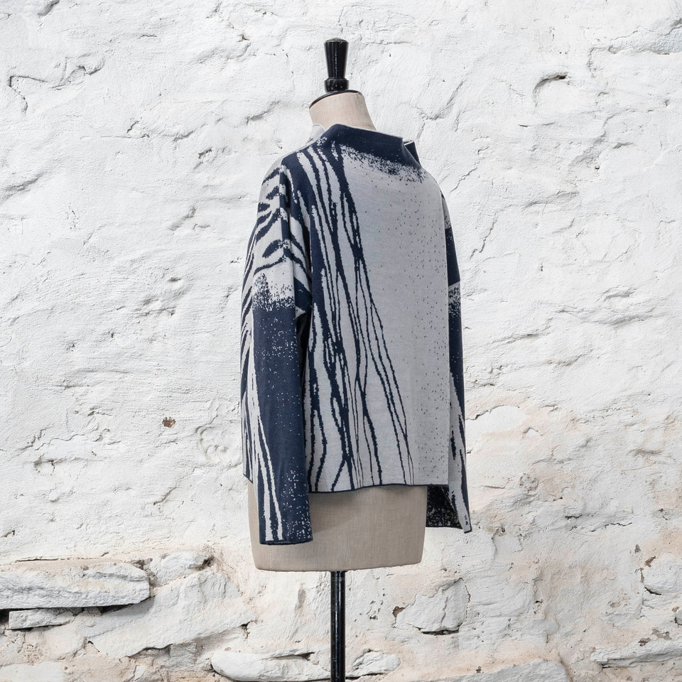 on a vintage mannequin against a rustic whitewashed wall, a finely knitted merino jumper in double faced jacquard. A linear design flows in waves down one side of the jumper - here on the back left shoulder in deep midnight blue against off white. There are areas of white with speckle and blue areas with white speckle. The jumper is shown from the left at a three quarter angle and has a stand up neckline
