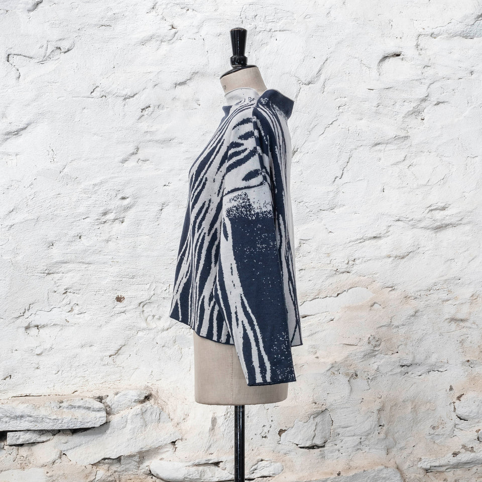on a vintage mannequin against a rustic whitewashed wall, a finely knitted merino jumper in double faced jacquard. A linear design flows in waves down one side of the jumper - here on the back left shoulder in deep midnight blue against off white. There are areas of white with speckle and blue areas with white speckle. The jumper is shown from the left and has a stand up neckline