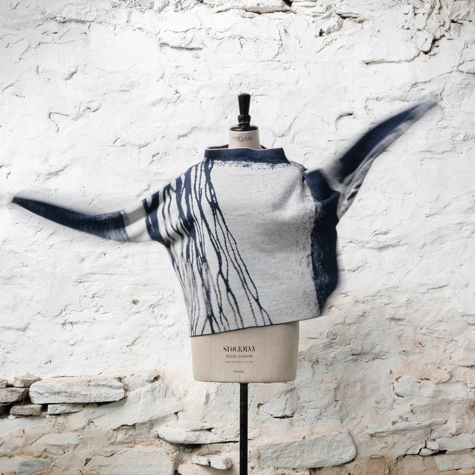 on a vintage mannequin against a rustic whitewashed wall, a finely knitted merino jumper in double faced jacquard. A linear design flows in waves down one side of the jumper - here on the front in a deep midnight blue against off white. other areas are just seen as the sleeves of the jumper fly up. the jumper has a stand up neckline.