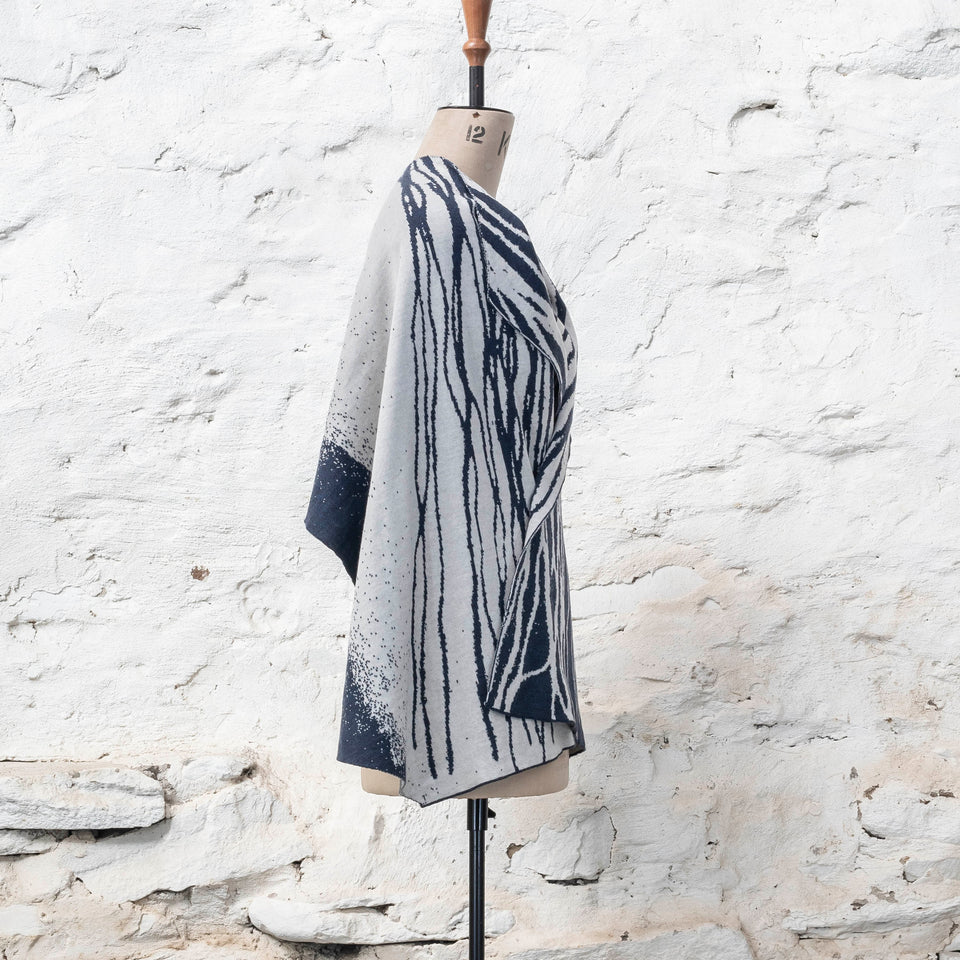 A midnight blue and off white knitted open cape in fine merino yarn. the pattern has fluid travelling likes either in blue on white or reversed out in white on blue. Shown from the right side.
