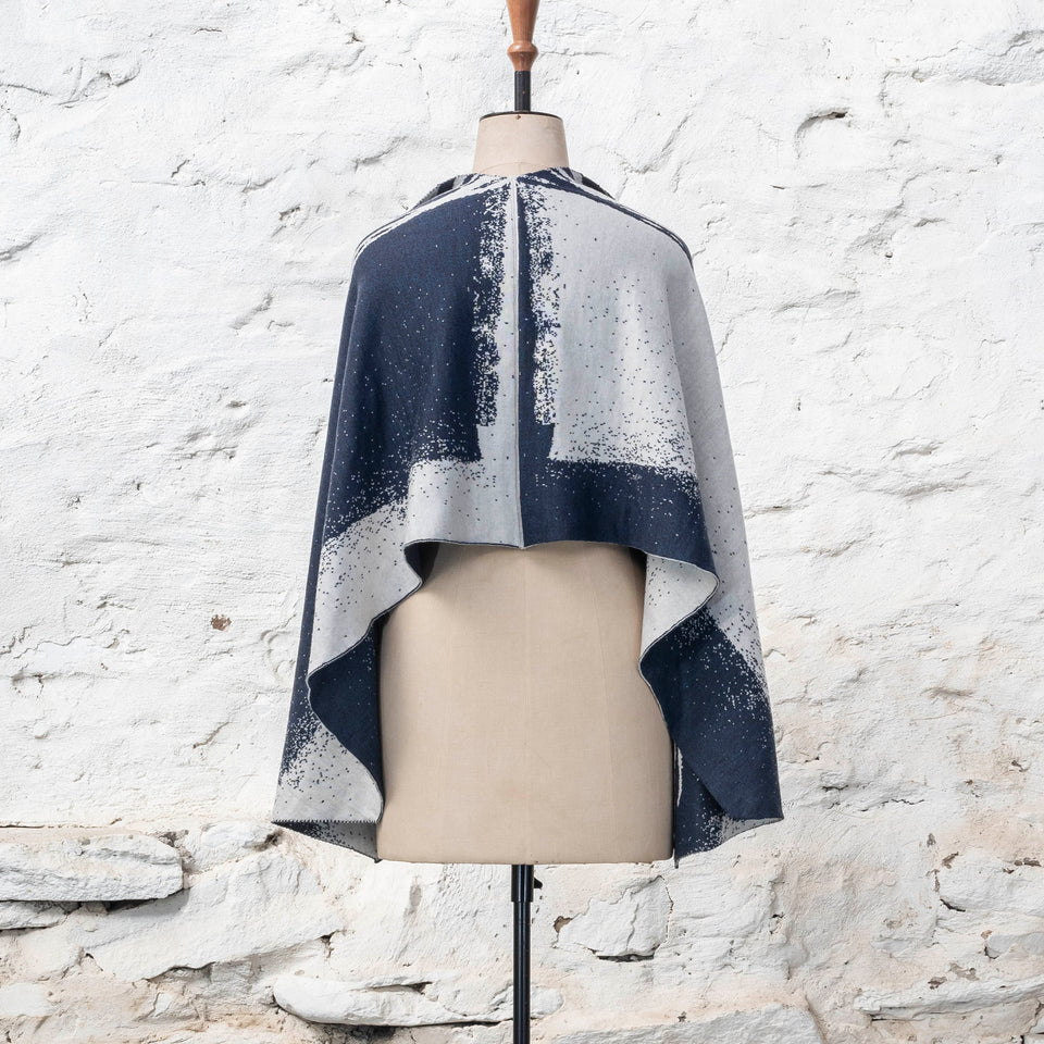 A midnight blue and off white knitted open cape in fine merino yarn. the pattern has fluid travelling likes either in blue on white or reversed out in white on blue. Shown from the back with the flat edge along the back