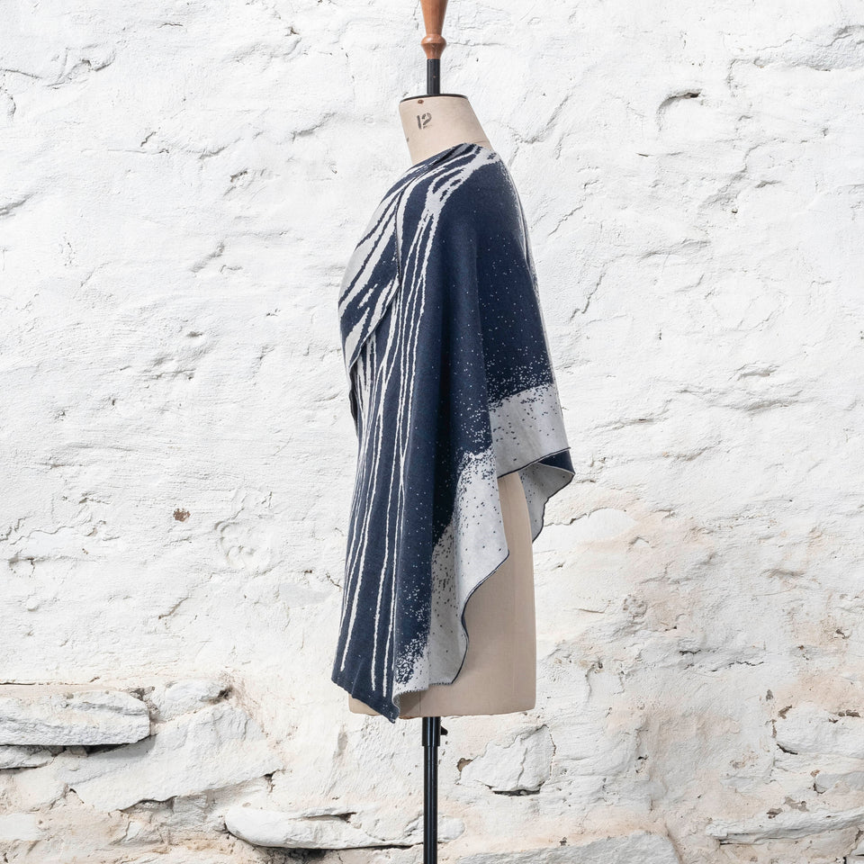 A midnight blue and off white knitted open cape in fine merino yarn. the pattern has fluid travelling likes either in blue on white or reversed out in white on blue. Shown from the left side