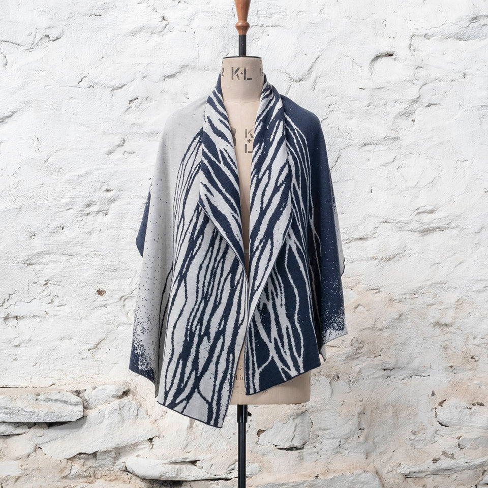 A midnight blue and off white knitted open cape in fine merino yarn. the pattern has fluid travelling likes either in blue on white or reversed out in white on blue. Arranged on a vintage mannequin with a cowl neck.