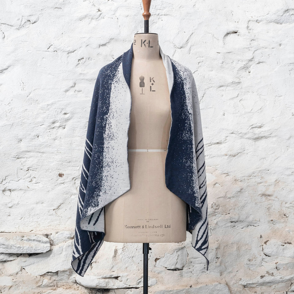 A midnight blue and off white knitted open cape in fine merino yarn. the pattern has fluid travelling likes either in blue on white or reversed out in white on blue. Shown from the front, open with plainer panels to the front.