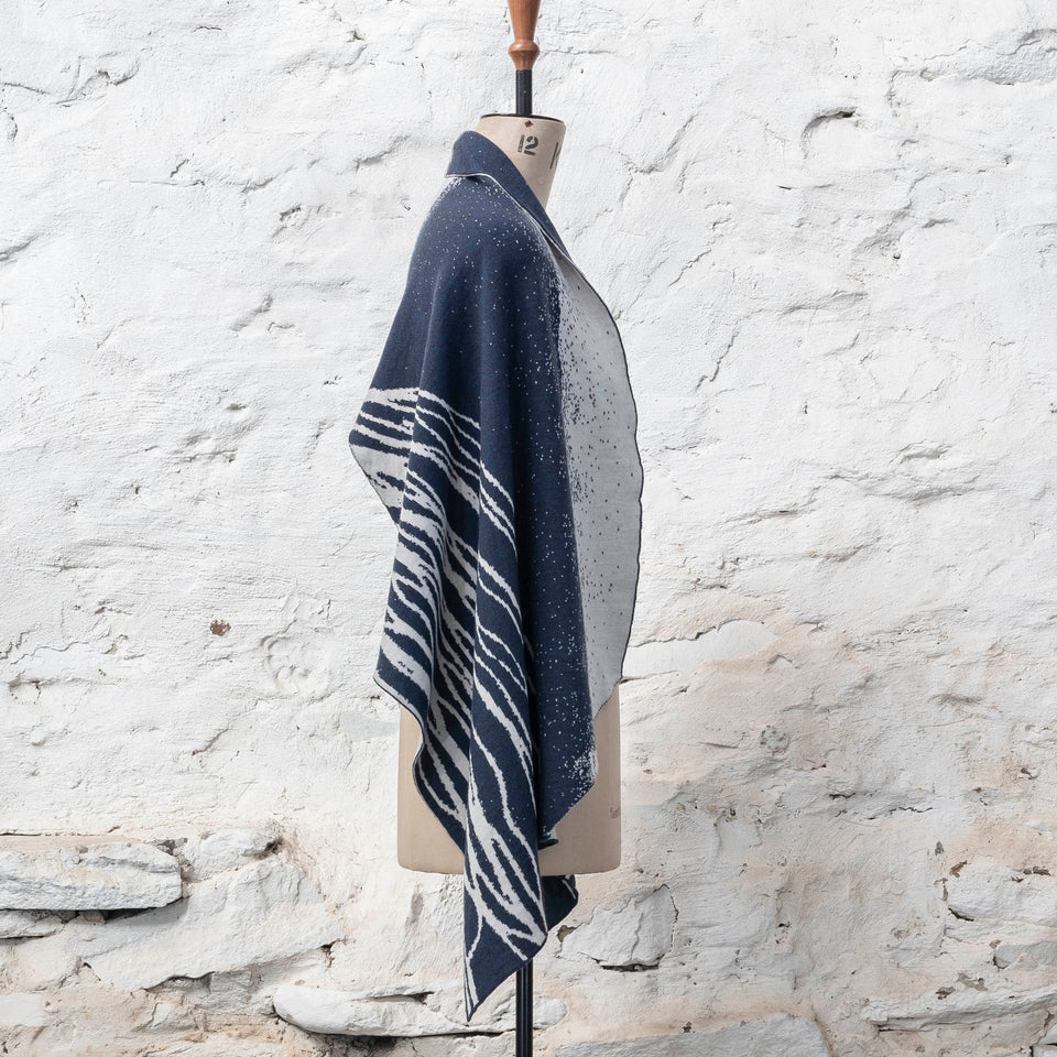 A midnight blue and off white knitted open cape in fine merino yarn. the pattern has fluid travelling likes either in blue on white or reversed out in white on blue. Shown from the right with plainer panels to the front