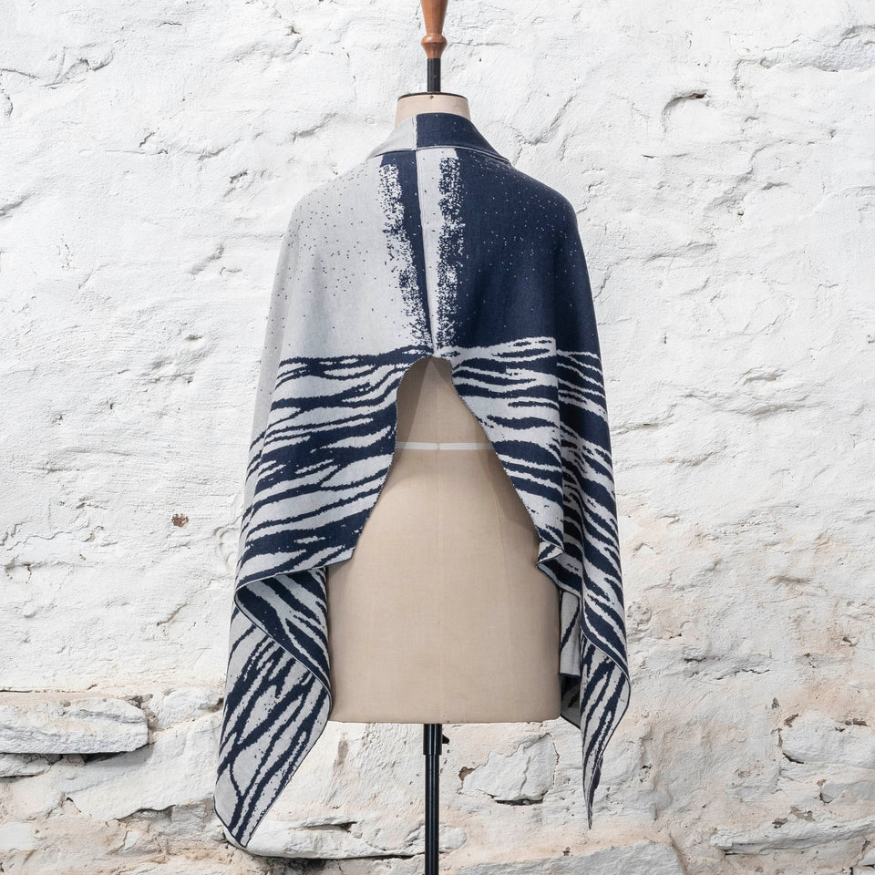 A midnight blue and off white knitted open cape in fine merino yarn. the pattern has fluid travelling likes either in blue on white or reversed out in white on blue. Shown from the back with the split at the back showing.